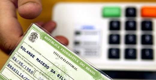 Homem segurando título eleitoral e leitor biométrico ao fundo.